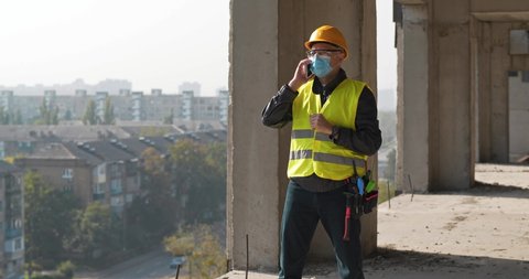 Workman in white hard hat and medical protective mask stands at construction site and speaks on smartphone. Cinema 4K 60fps panoramic video
