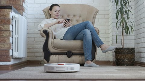 House cleaning robot. Woman in a chair with a smartphone cleans the carpet with a smart vacuum cleaner.