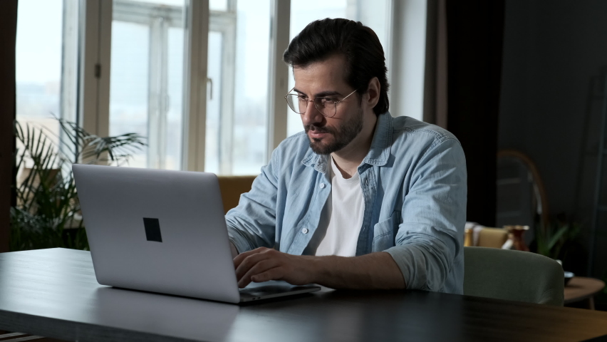 Smart Man in Glasses, Blue Shirt and White t-Shirt Working on a Computer laptop Online, Sitting at a Table at Home in the Evening in a Dark Room Royalty-Free Stock Footage #1063298473
