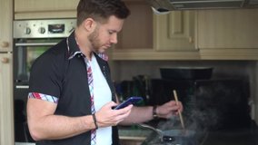 Young Man Cooking Meal Whilst Looking At Mobile Phone