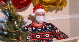 Handsome guy in christmas sweater, medical mask and santa hat talking on the phone while sitting on his desk with laptop on Christmas Eve with 2021 balloons on the background. Celebrating at home.