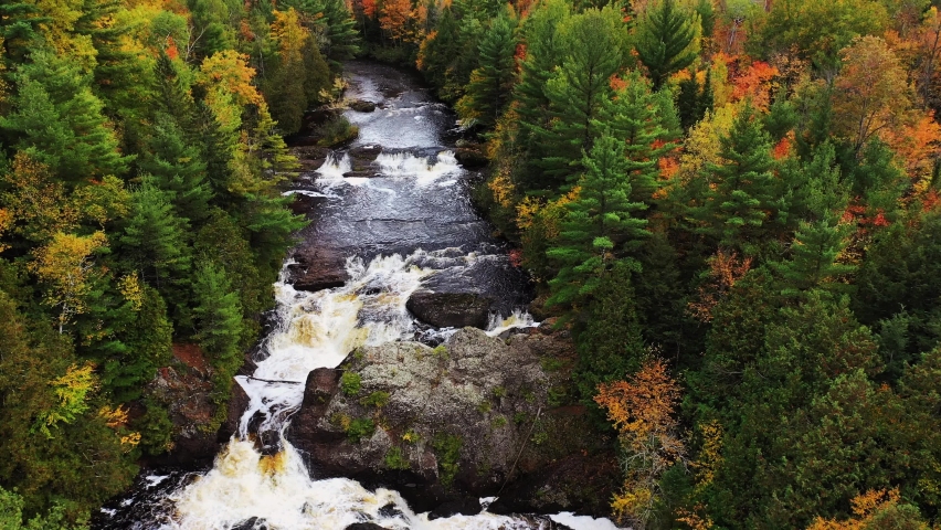 Beautiful autumn aerial flying over Upper Potato Falls with heavy water flow cascading over large rock formations with yellow orange and green deciduous and evergreen trees lining the river banks.