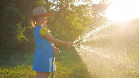 Funny little girl in hat playing with garden hose in sunny backyard. Adorable little girl playing with a garden hose on hot and sunny summer evening. Summer outdoors activity for kids.