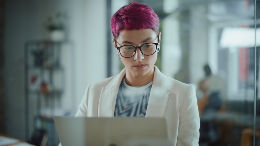 Modern Office: Portrait of Beautiful Authentic Specialist with Short Pink Hair Standing, Holding Laptop Computer, Looking at Camera, Smiling Charmingly. Working on Design, Data Analysis,Plan Strategy Royalty-Free Stock Footage #1063309399