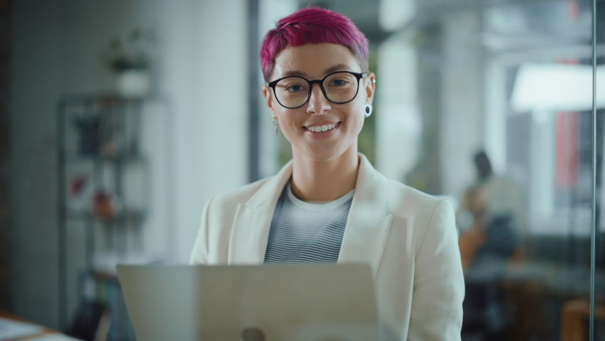 Modern Office: Portrait of Beautiful Authentic Specialist with Short Pink Hair Standing, Holding Laptop Computer, Looking at Camera, Smiling Charmingly. Working on Design, Data Analysis,Plan Strategy | Shutterstock HD Video #1063309399