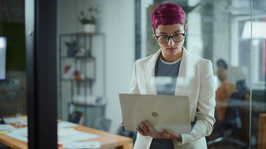 Modern Office: Portrait of Beautiful Authentic Specialist with Short Pink Hair Standing, Holding Laptop Computer, Looking at Camera, Smiling Charmingly. Working on Design, Data Analysis,Plan Strategy | Shutterstock HD Video #1063309405