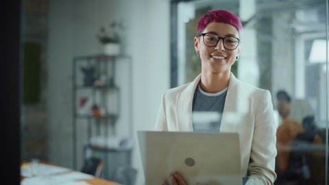 Modern Office: Portrait of Beautiful Authentic Specialist with Short Pink Hair Standing, Holding Laptop Computer, Looking at Camera, Smiling Charmingly. Working on Design, Data Analysis,Plan Strategy