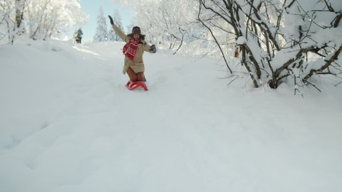 Young man riding sled on his knees down the hill and then falling down in snow while having fun with friends in forest on winter day