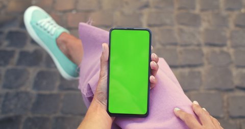 Crop view of female person waving while having video call on smartphone with mockup screen and sitting at street. Concept of chroma key and greenscreen.Paving stones background