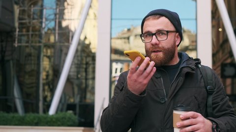 Bearded man making voice message or using virtual assistant app and laughing while looking at screen. Handsome guy in glasses talking and using smartphone while walking at street