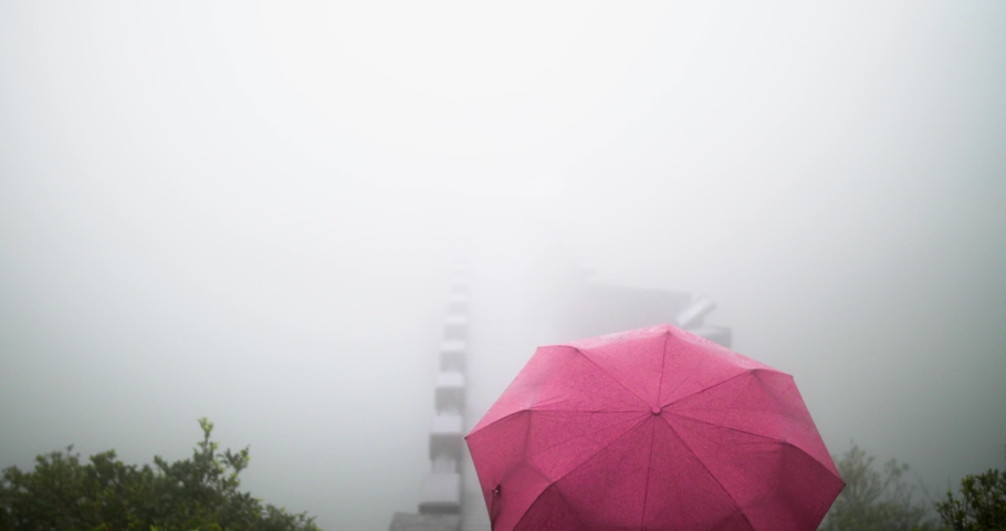 Girl with red umbrella walking across a bridge into dense white fog and clouds. Mysterious young traveler exploring nature during rainy day. Yangmingshan national park, Taiwan, Republic of China. 4K.