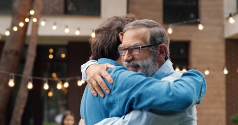 Cheerful happy Caucasian man meeting with old father and hugging at back yard of house. Family dinner outdoors on background. Senior dad with adult son in hugs. Generations. Two men embracing.