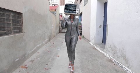 Footage of woman with television covering face and head walking down an alleyway