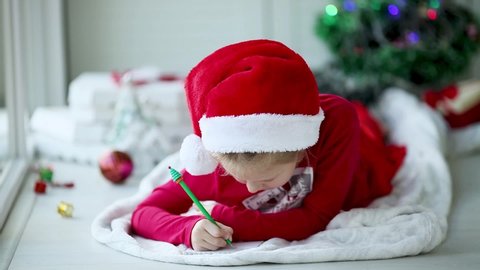 A smiling girl in a Santa hat writes a letter, dreams of gifts to Santa Claus. Child lying on a blanket on the floor of a house with christmas lights