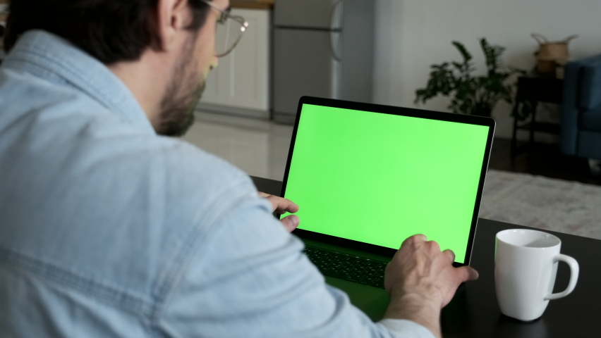 Over the Shoulder Camera Shot of Man at Home Sitting on a Couch Working on Green Mock-up Screen Laptop Computer Online  Royalty-Free Stock Footage #1063324369
