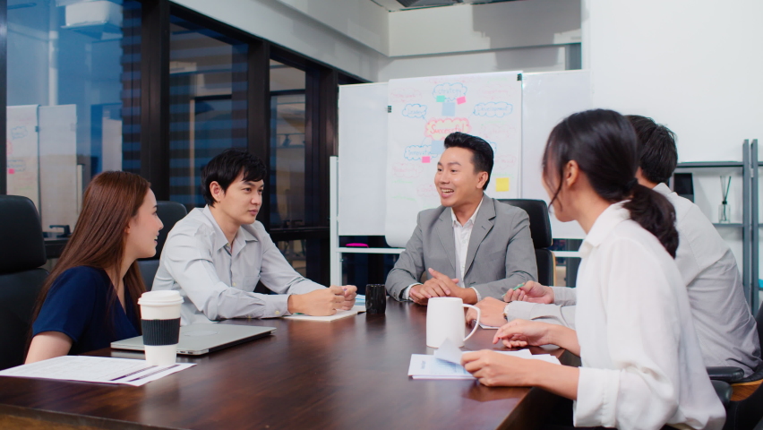 Group of happy Asian business people discuss together in team brainstorm meeting and clap hand, work late night in office. Corporate business, coworker teamwork, or project success celebration concept Royalty-Free Stock Footage #1063326817