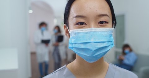 Close up portrait of Asian happy young beautiful female hospital practitioner wearing mask while standing in clinic hall and smiling at camera. Coronavirus pandemic. Healthcare worker. Covid-19