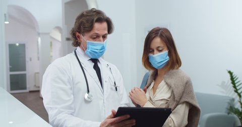 Close up portrait of male physician standing in clinic and talking to female pretty patient in medical mask and browsing on tablet. Multi-ethnic healthcare workers speak on background Covid-19 concept