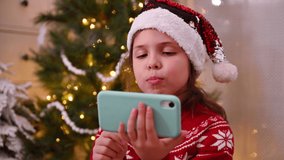 Little girl in a red sweater with a phone near the Christmas tree in a sweet home. A child in a Santa hat plays online on a smartphone. 