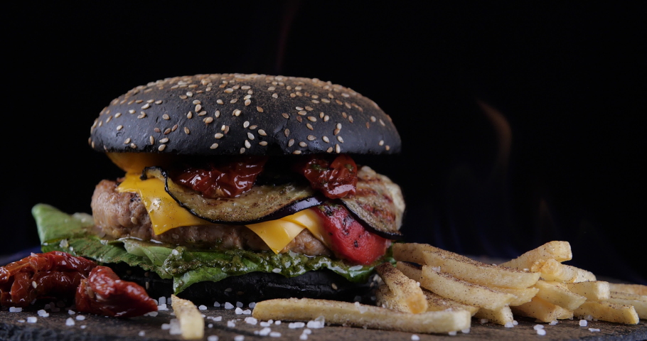 Black burger with meat and vegetables with fries lie on table against background of fire. Chisburger with salad tomatoes onion sauce and beef and dark bread bun on grill surrounded by flames hot dish