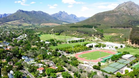 Aerial drone rotating, university college sports athletics stadium, track and field, tennis courts, mountains, trees and neighbourhood in background, Stellenbosch, Coetzenburg