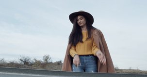 a young and beautiful girl dressed stylishly with a hat on her head and a brown coat. A smiling woman walks on a sunny day on the road. Slow motion video.