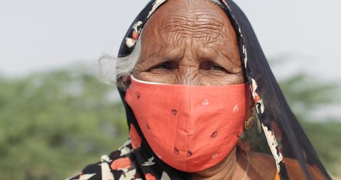 Static CU close-up of elderly Indian woman outdoors during the coronavirus global pandemic wearing a face mask on terrace balcony looking directly at camera pov and speaking from behind, slow-motion