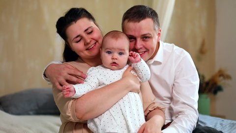 Portrait of joyful young family with little baby girl sitting on bed with sunny home interior on background. Weekend together. Happy mother and father enjoying parenting. Excited female child.