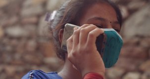 Indian teenage girl wearing a matching colored face mask outdoors in rural streets using a smart phone mobile touchscreen technology device to type text message and voice video call to communicate 