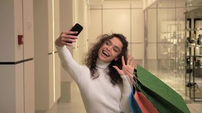 Charming girl with curly hair walks around a shopping mall while having a video call. Young beautiful woman holds colorful shopping bags in hand