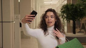 Beautiful girl with curly hair walks around a shopping mall while having a video call. Young woman talks with her friend using smartphone holds colorful shopping bags in hand