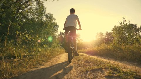 Happy kid riding bike in park at sunset. Cycling along the path in woods in the rays of beautiful sunset. Active rest of child. Healthy lifestyle, child sports. Child riding a bike in a forest park
