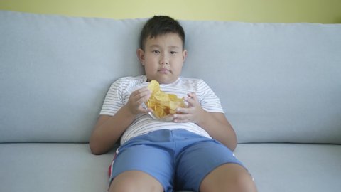 Overweight Asian boy eating unhealthy potato chips while sitting on sofa