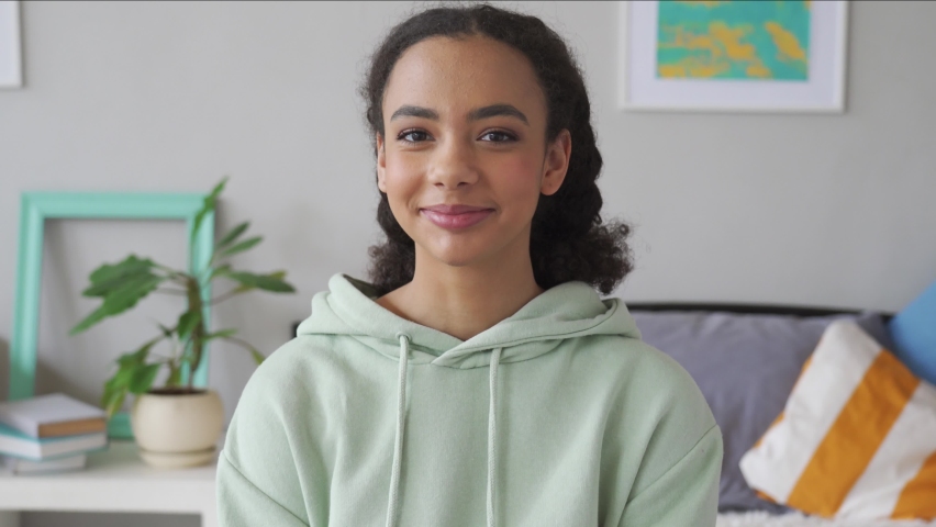 Happy african american teen girl looking at camera at home. Cheerful generation z mixed race pretty teenager smiling while sitting on bed in bedroom casual interior, headshot portrait. Royalty-Free Stock Footage #1063356874