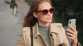 Portrait of pretty girl wearing sunglasses and trench coat talking with friend by video call on smartphone outdoor