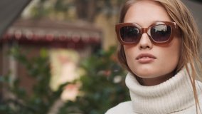 Beautiful stylish girl in sunglasses dressed in white sweater posing on camera on city street