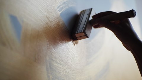 Close-up of artist holding paint wide brush and drawing oil painting at the studio. Unusual new abstract painting, brown, gold line on light canvas. One example of a painting technique.