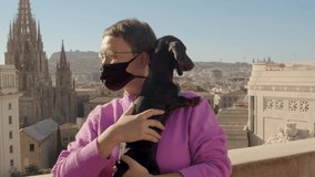 Woman with short haircut in a purple sweater and black anti COVID-19 medical mask is holding a dachshund dog puppy. Montjuic mountain, Cathedral, Barcelona, Spain. Panned video 4k high quality footage
