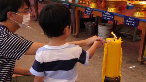 4K Buddhist little child boy lighting incense joss stick from big candle with father in local temple in countryside. Translation:"candle for remove bad luck" "increase fortune" "40 baht" 