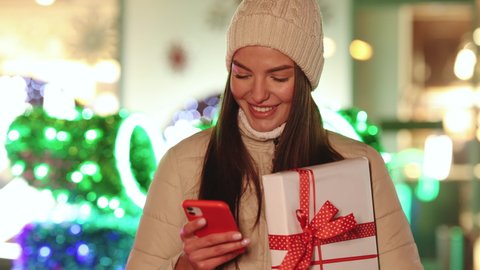 Portrait of happy young Caucasian woman standing outdoor with christmas gift and texting on smartphone. Cheerful female with xmas present on decorated street tapping on cellphone. 4k Slow motion
