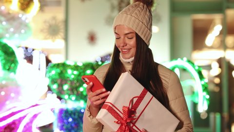 Portrait of young woman stand outdoors with christmas present texting on smartphone. Gift box in city typing use phone. Lights around. Smiling feel happy. Gift Box.