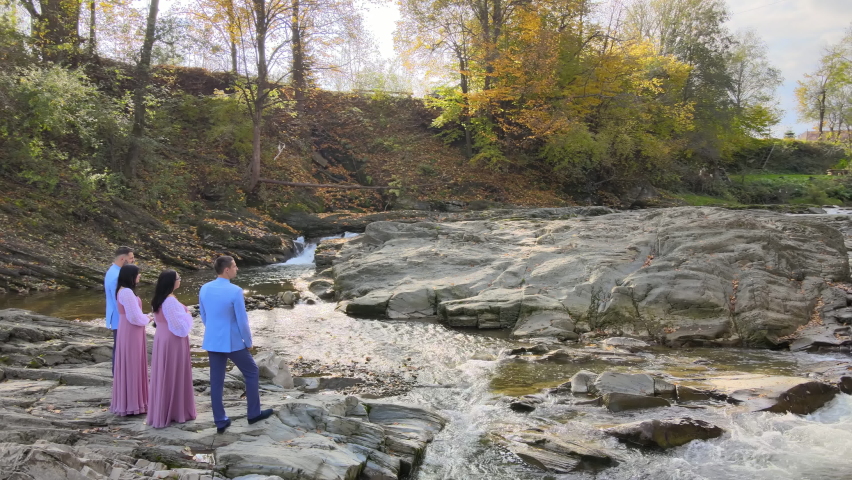 Group of four people, two men in suits and two women and dresses standing near small mountain river in autumn warm day.