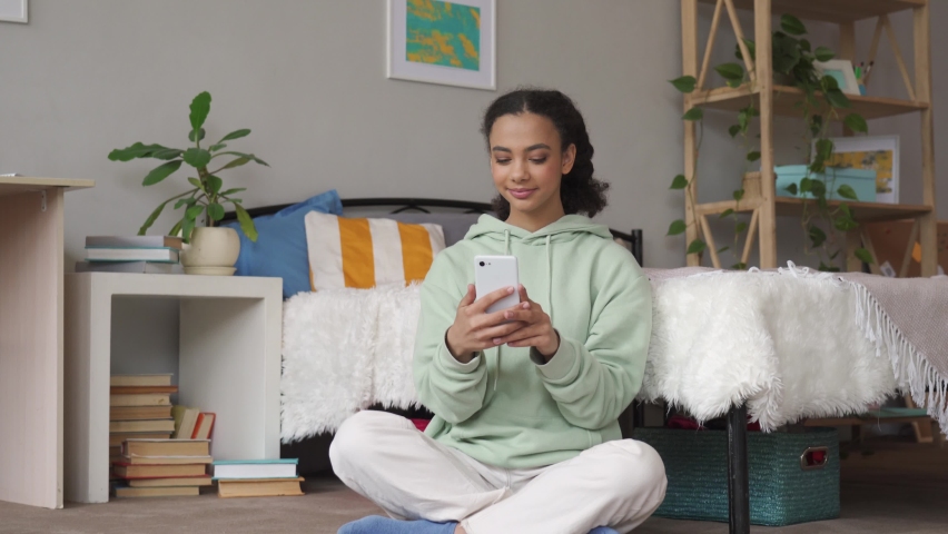 Happy gen z african american teen girl influencer blogger using mobile cell phone, taking selfie, posting in social media apps, chatting with friends online on smartphone sitting in bedroom at home.
