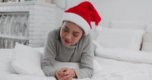 Lovely young asian woman lying in bed wear Christmas hat talking to camera say Merry Christmas to friend lovely girl making video call at home