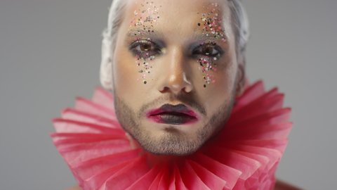 Close up shot of young man with red ruff around his neck and full face of theatrical make-up with glitter looking at camera