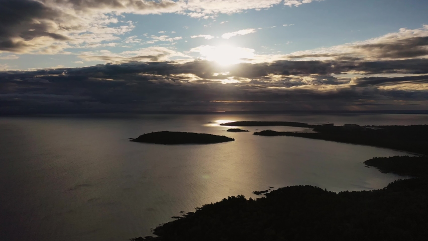 Beautiful panning down sunrise aerial above Sugarloaf Mountain looking towards Presque Isle peninsula and islands in Lake Superior with the sun shining between clouds with fall foliage on trees below.