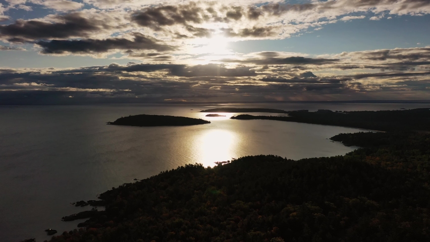 Beautiful panning across sunrise aerial above Sugarloaf Mountain looking towards Presque Isle peninsula and islands in Lake Superior with the sun shining between clouds and reflecting off of the water