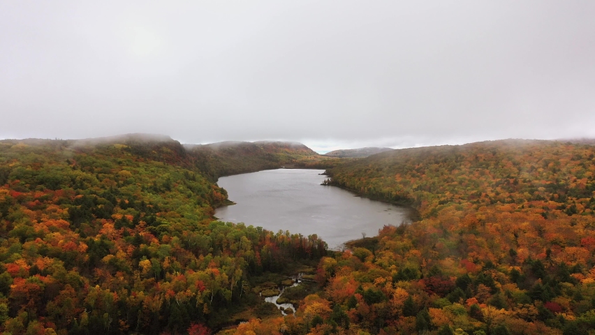 Beautiful panning down travel aerial of Lake of the Clouds and the pedestrian bridge over Carp River on an overcast morning with colorful autumn foliage blanketing the hills and valley below.