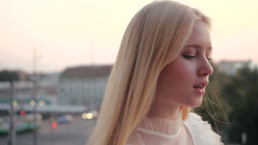 Close-up view of cute beautiful blonde girl in a light white dress walking along the evening city center and fluffing her silky hair in the wind against a blurry background of trees and cars driving. Royalty-Free Stock Footage #1063370686