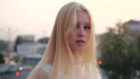 Close-up view of cute beautiful blonde girl in a light white dress walking along the evening city center and fluffing her silky hair in the wind against a blurry background of trees and cars driving.